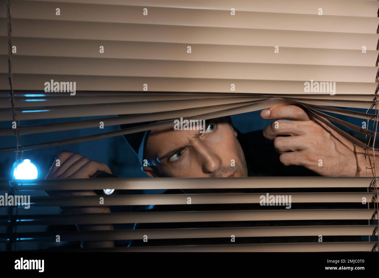 Male security guard with flashlight looking through window blinds in