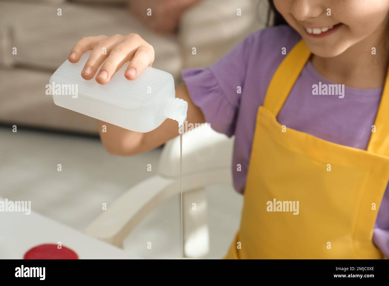Little girl pouring glue into bowl at table in room, closeup. DIY slime