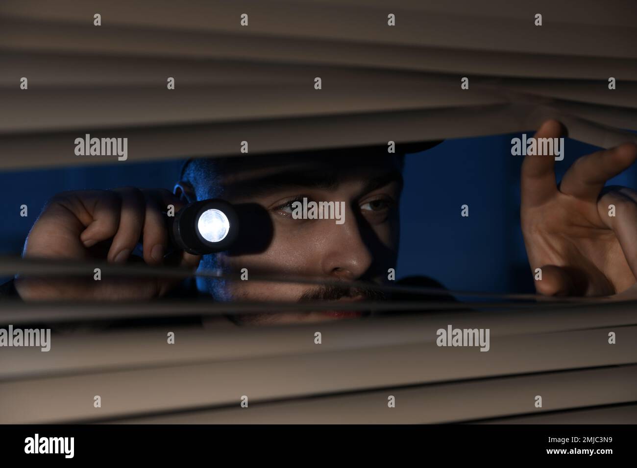 Male security guard with flashlight looking through window blinds in