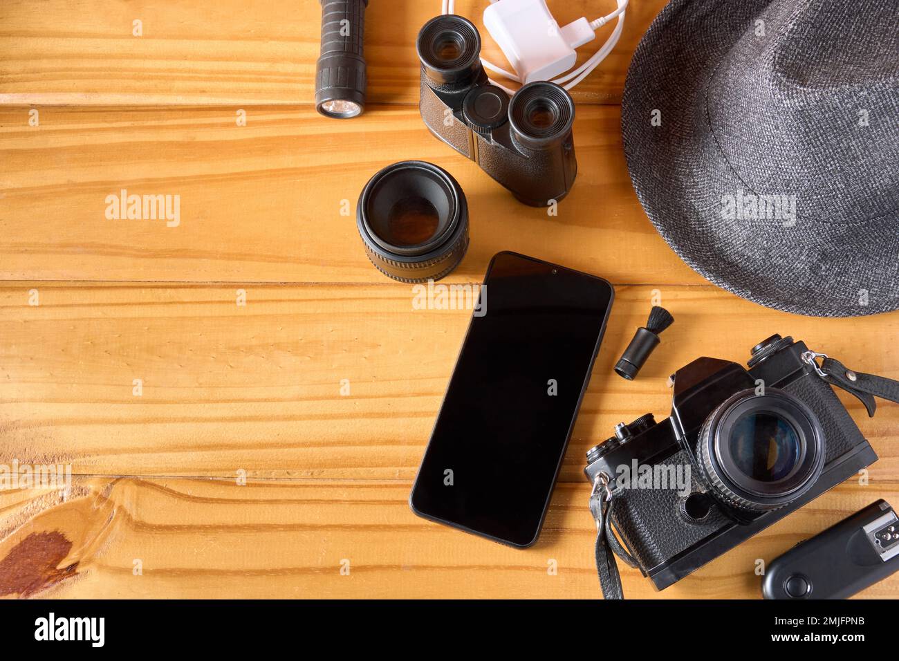 Top view of travel accessories on wooden table with mobile phone, camera and lens, hat, binoculars, flashlight, charger, cleaning brush and transmitte Stock Photo