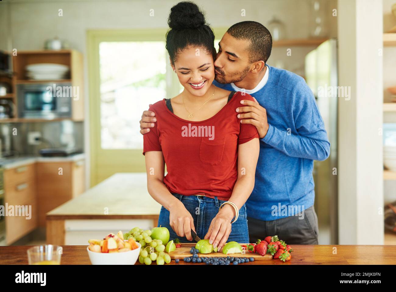 That looks good. a handsome young man kissing his wife while she makes a fruit salad in the kitchen. Stock Photo