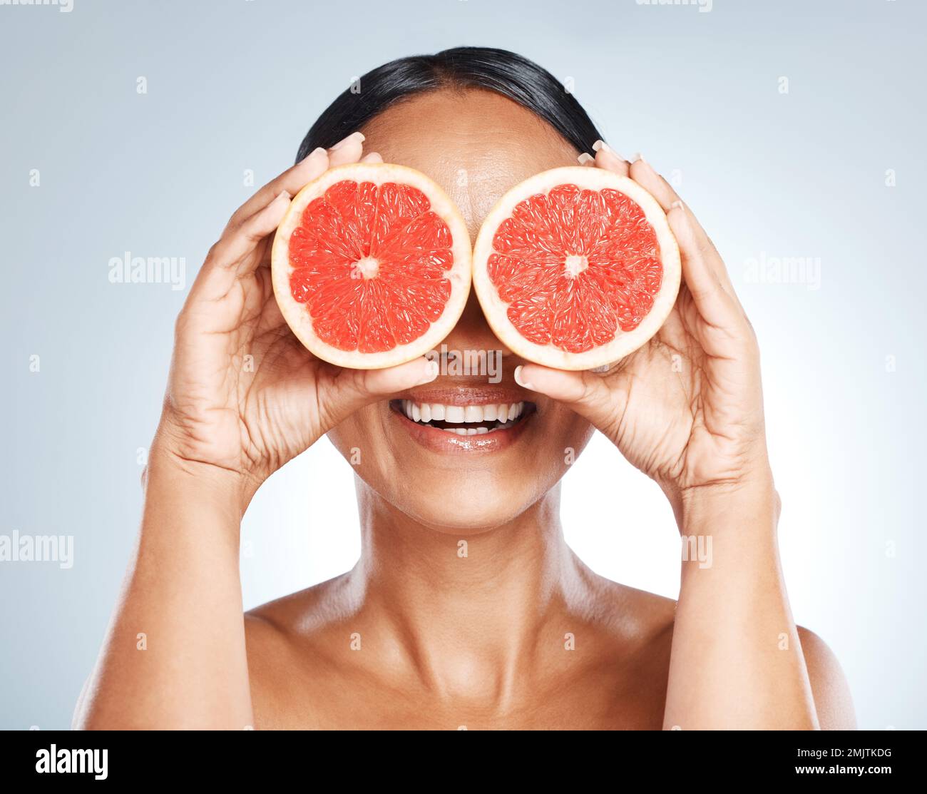 Funny, fruit and woman use grapefruit for a joke covering her eyes isolated against a studio blue background. Health, beauty and skincare model Stock Photo