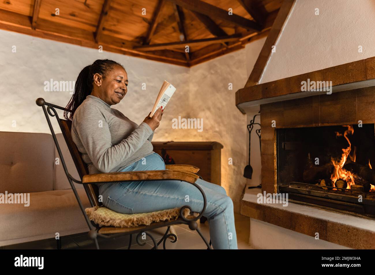 Senior African woman reading book while warming in front of fireplace in her house Stock Photo
