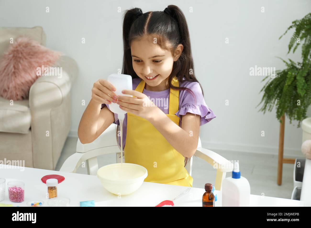 Cute little girl pouring glue into bowl at table in room. DIY slime toy