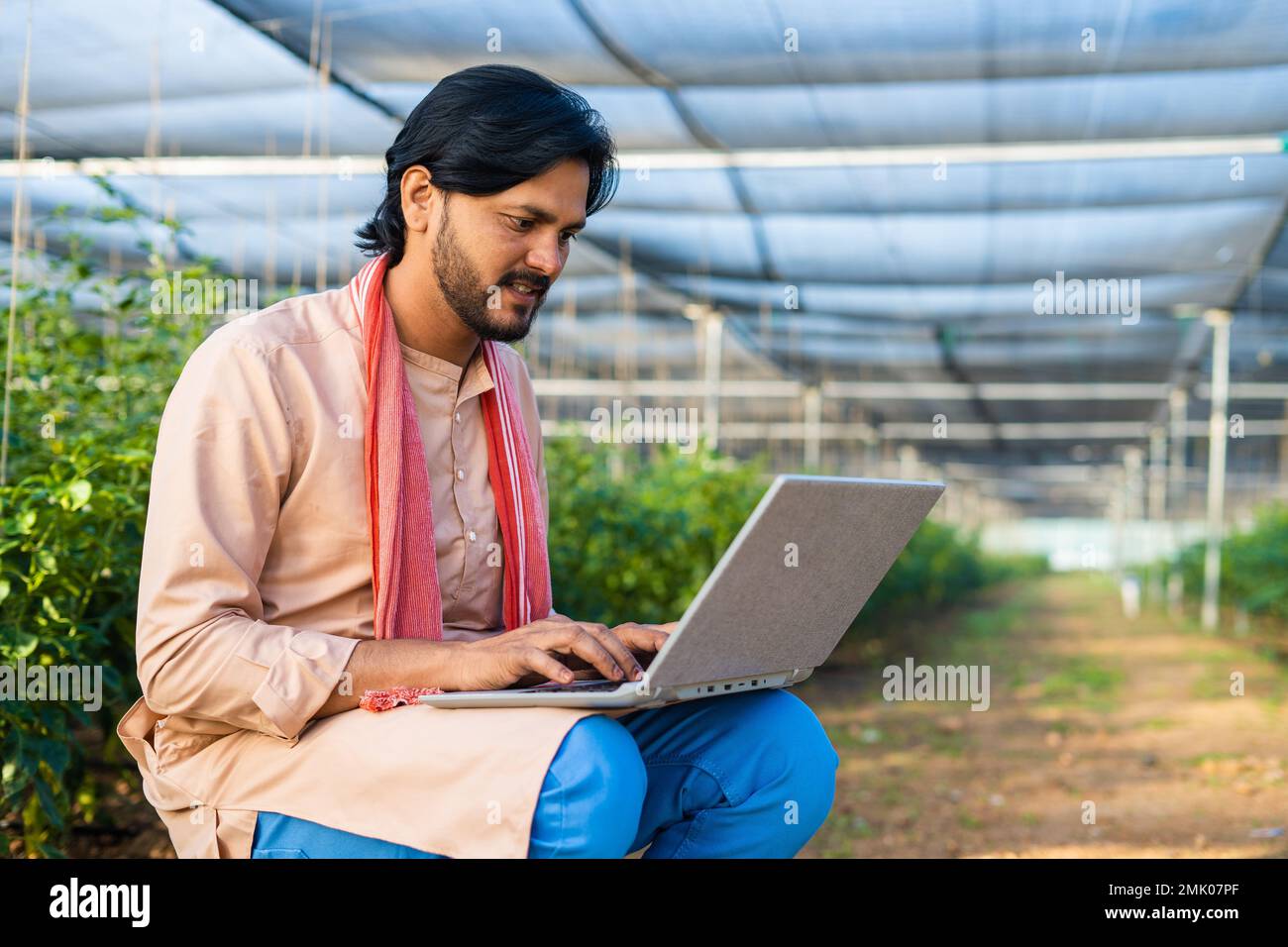 Happy young farmer busy working on laptop while sitting at greenhouse - concept of modern farming, technology and development or growth. Stock Photo