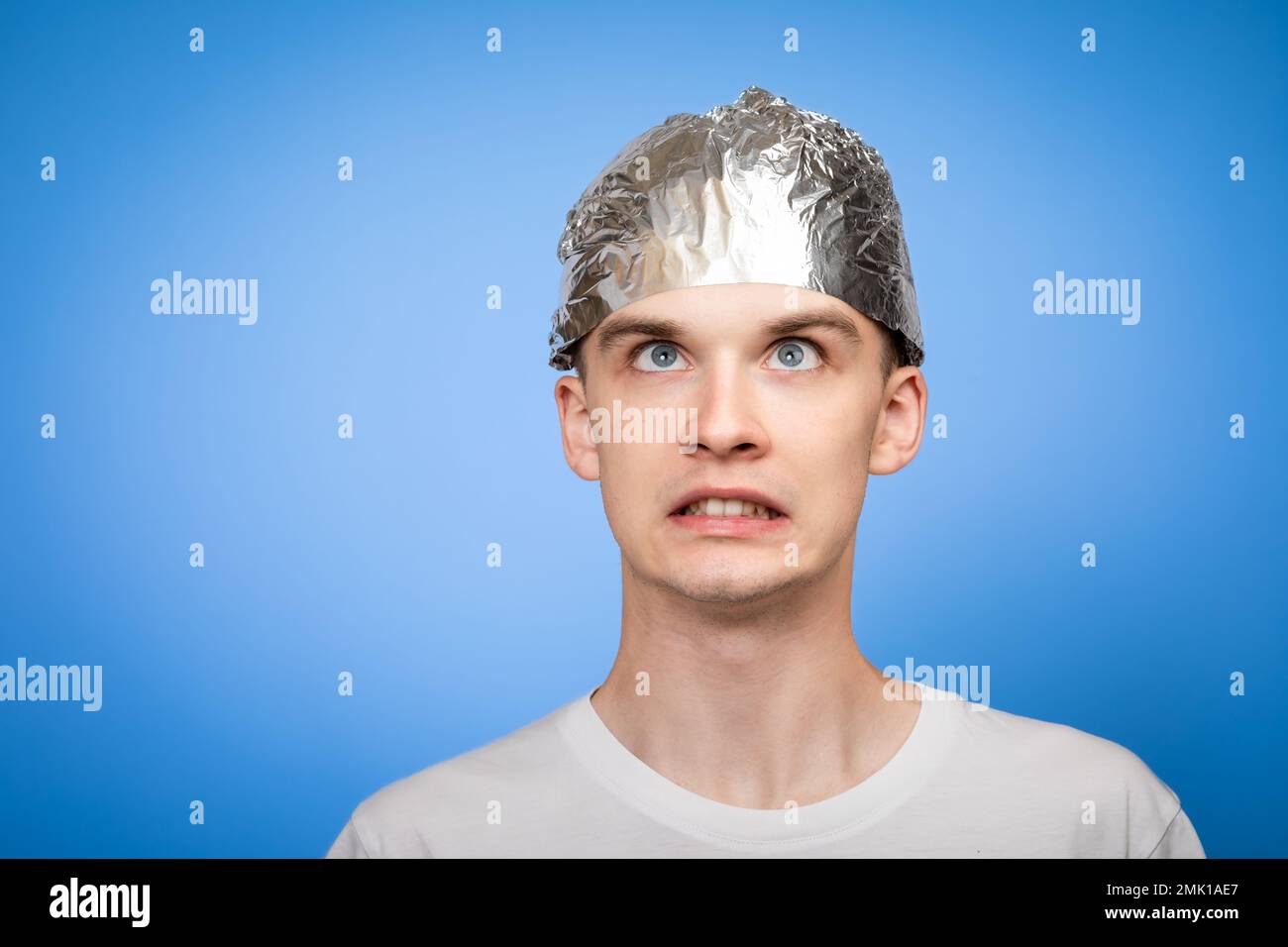 Portrait of anxious young man wearing tin foil hat looking up in panic. Conspiracy theories and paranoya concept. Studio shot on blue background Stock Photo