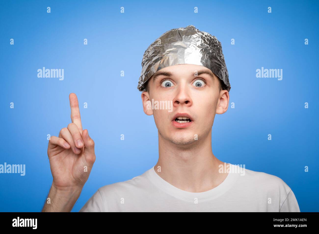 Portrait of anxious young man wearing tin foil hat pointing his finger up. Conspiracy theories and paranoya concept. Studio shot on blue background Stock Photo