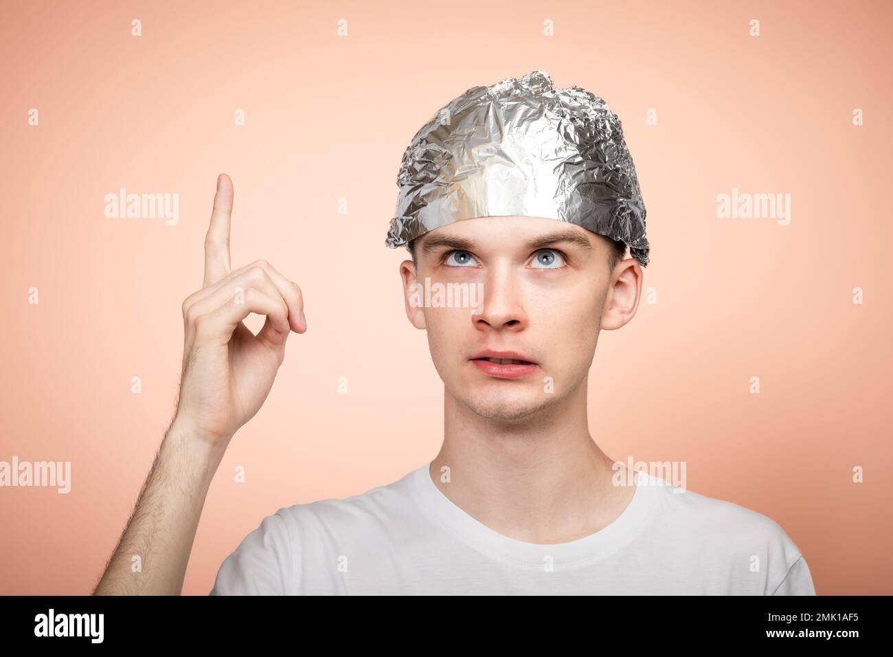 Portrait of anxious young man wearing tin foil hat pointing his finger and looking up. Conspiracy theories and paranoya concept. Studio shot on salmon Stock Photo