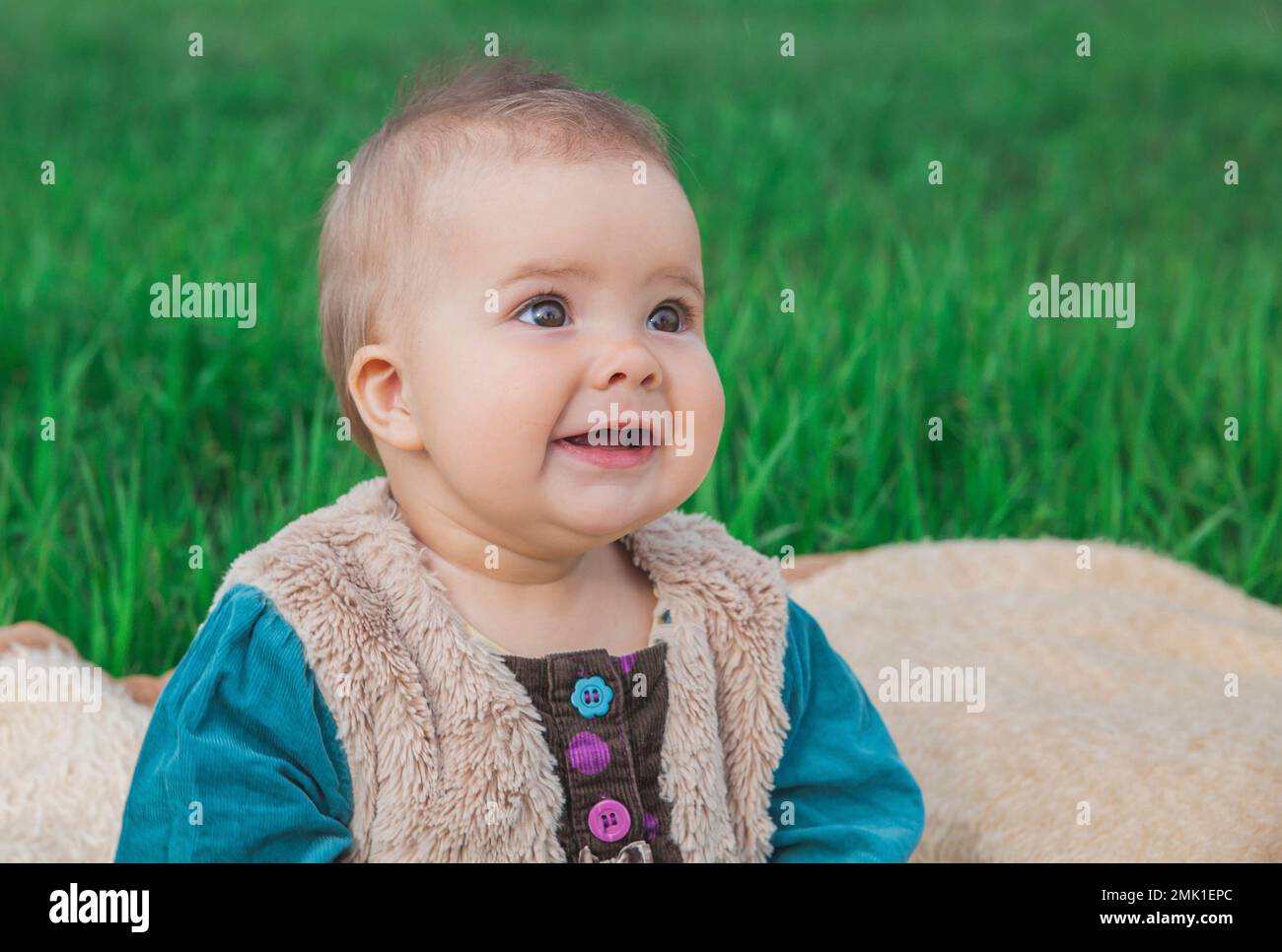 toothless baby in a multi-colored dress sitting on a bedspread on lawn Stock Photo