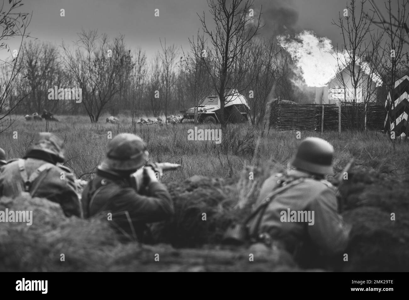 Re-enactors Armed Rifles And Dressed As World War Ii German Wehrmacht Infantry Soldiers Fighting Defensively In Trench. Fight Against Combat Vehicle Stock Photo