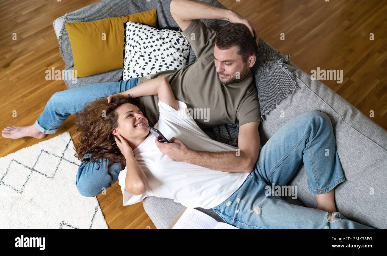 Couple lying on sofa in living room resting using mobile phone smiling, enjoying time together after work. Stock Photo
