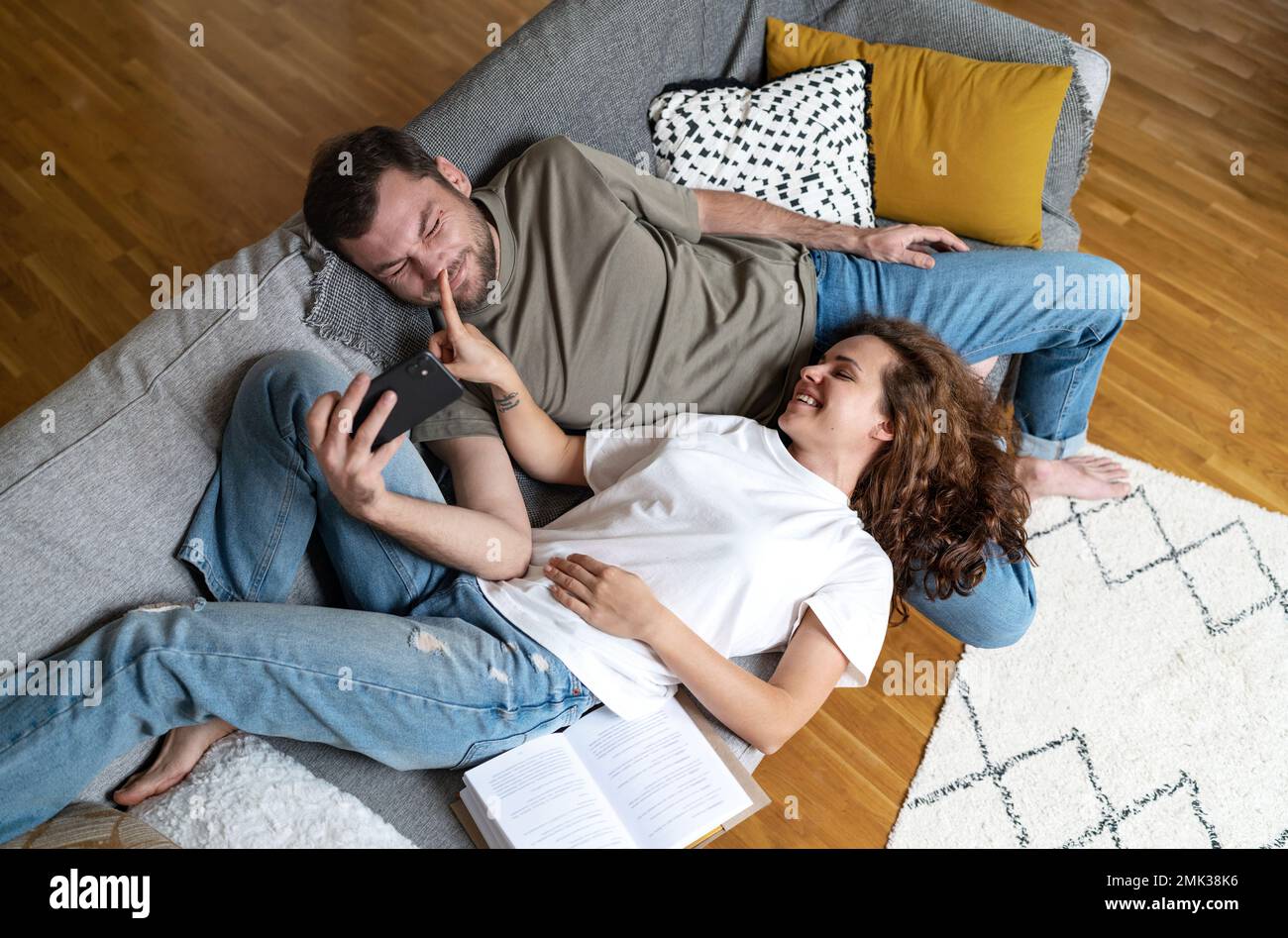 Playful couple lying down on sofa at home man using smart phone girlfriend touching boyfriends nose with her finger. Stock Photo
