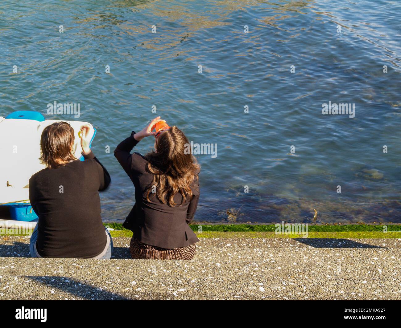 Wellington New Zealand - October 2 2010; Two young women sitting at edge of bay drinking on sunny day. Stock Photo