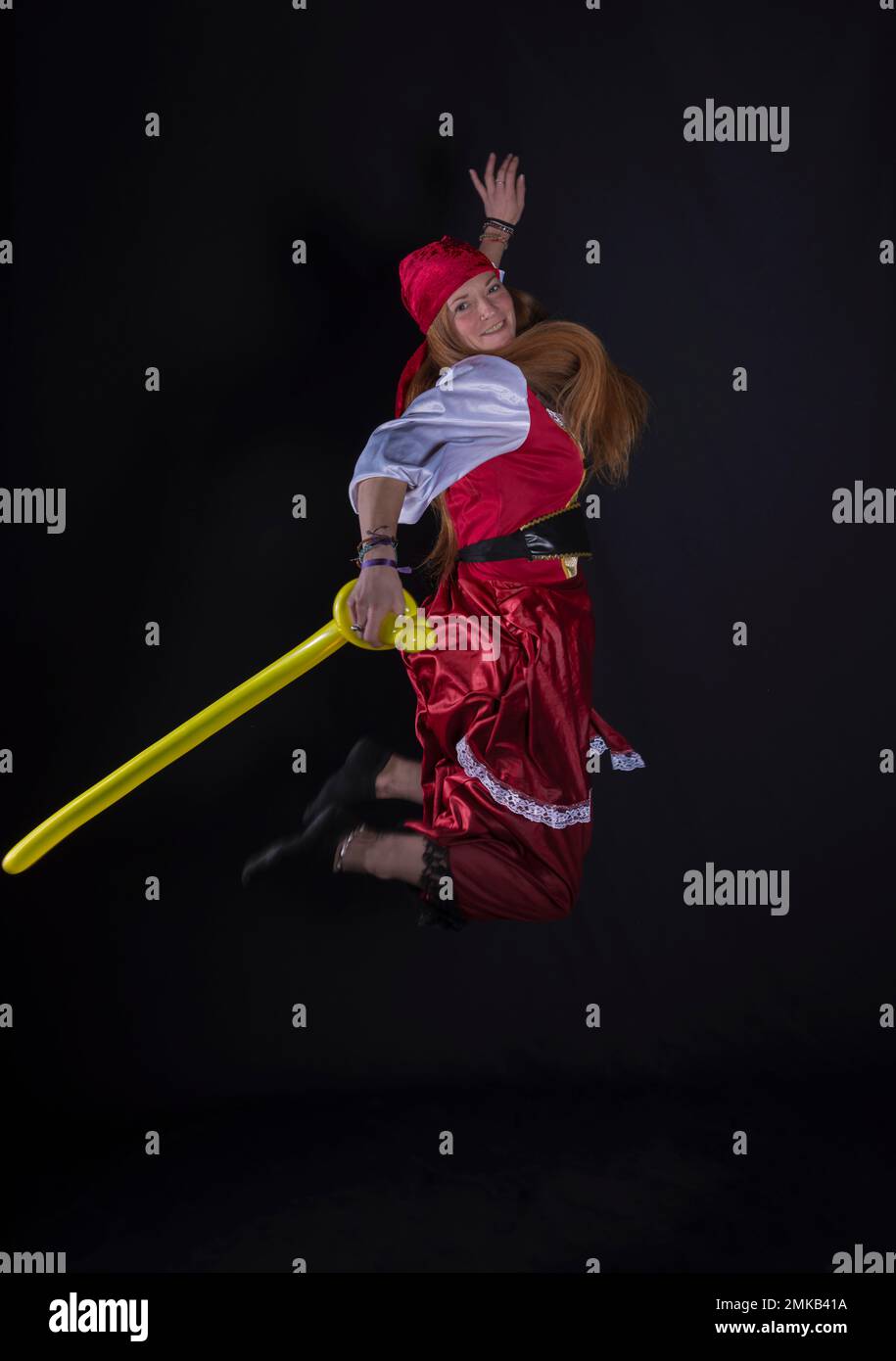 woman with long hair dressed as a pirate in a red suit and with a yellow balloon in the shape of a sword in her hand and on a black background photogr Stock Photo
