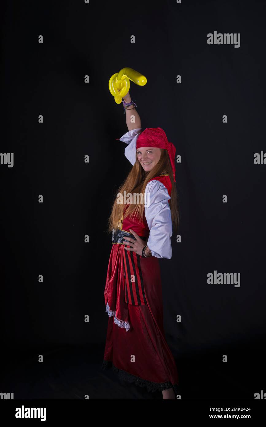 woman with long hair dressed as a pirate in a red suit and with a yellow balloon in the shape of a sword in her hand and on a black background photogr Stock Photo