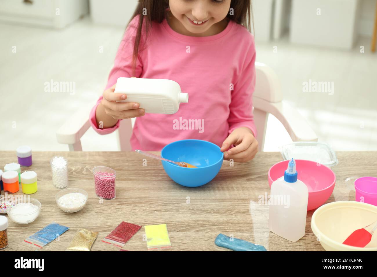 Little girl pouring glue into bowl at table in room, closeup. DIY slime