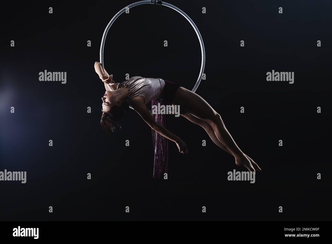 Young woman performing acrobatic element on aerial ring against dark ...