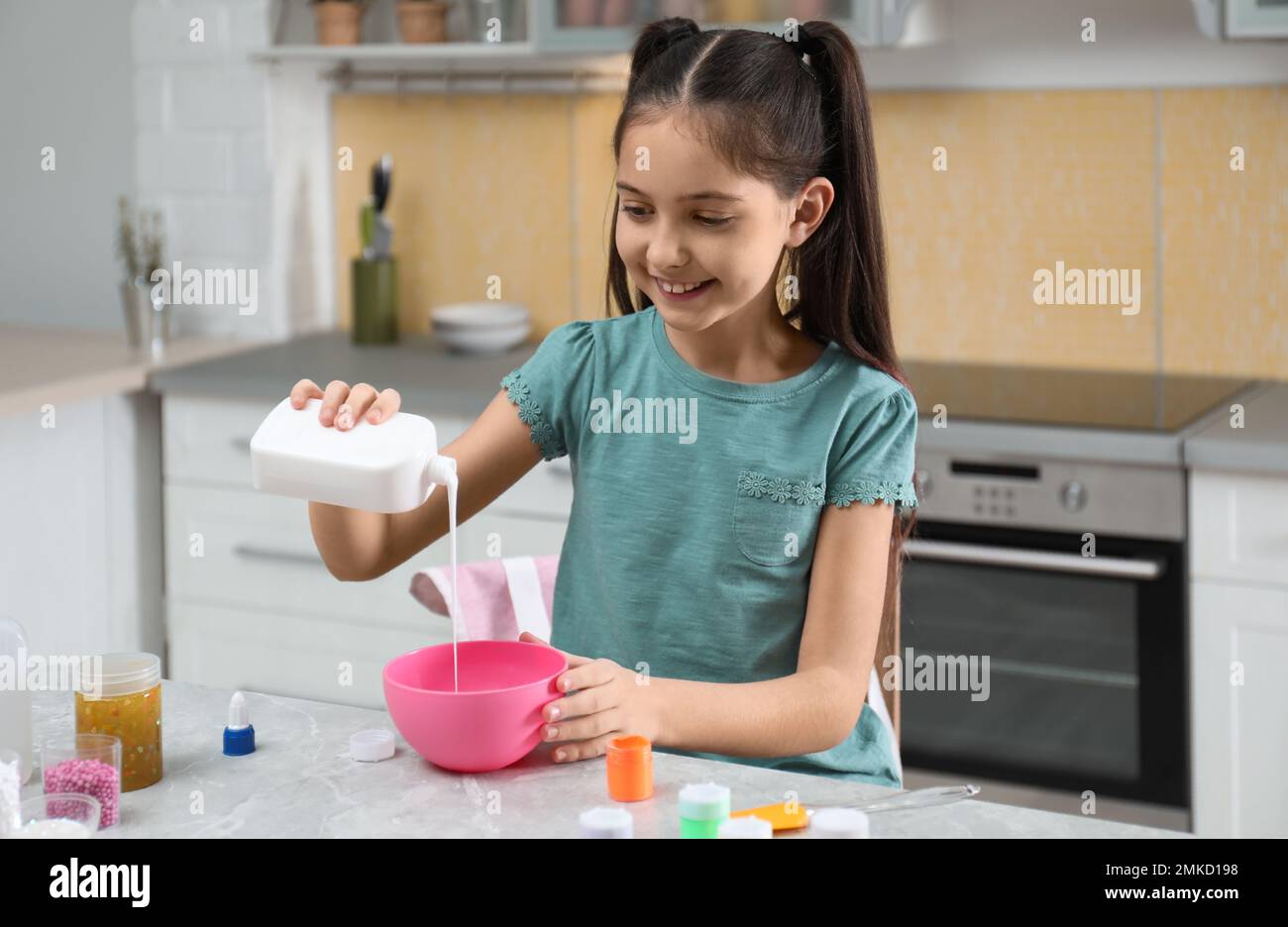 Cute little girl pouring glue into bowl at table in kitchen. DIY slime
