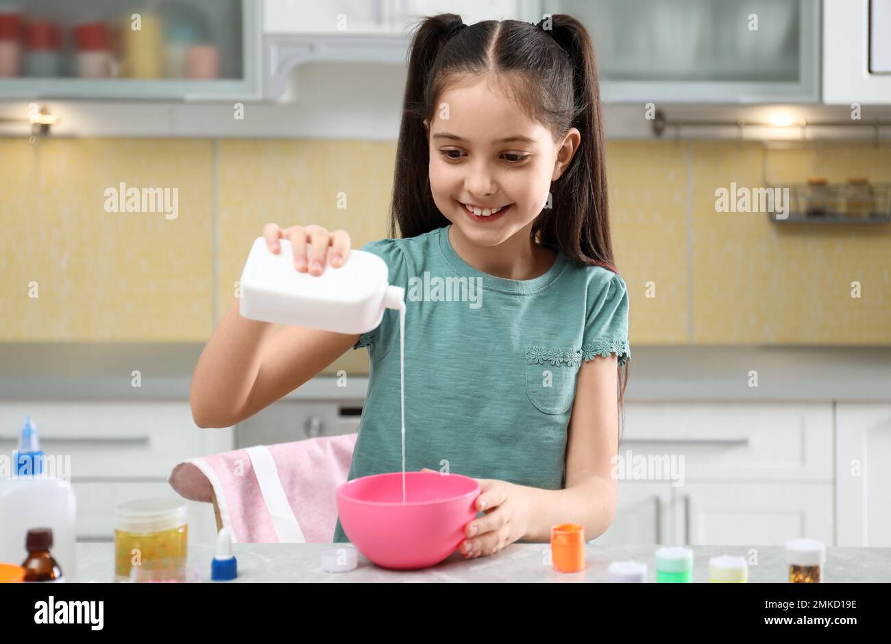 Cute little girl pouring glue into bowl at table in kitchen. DIY slime