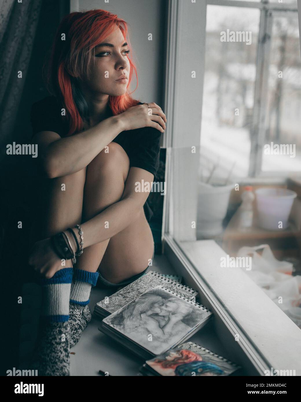 Young redhead woman artist is sitting on the window with her paintings and paints Stock Photo