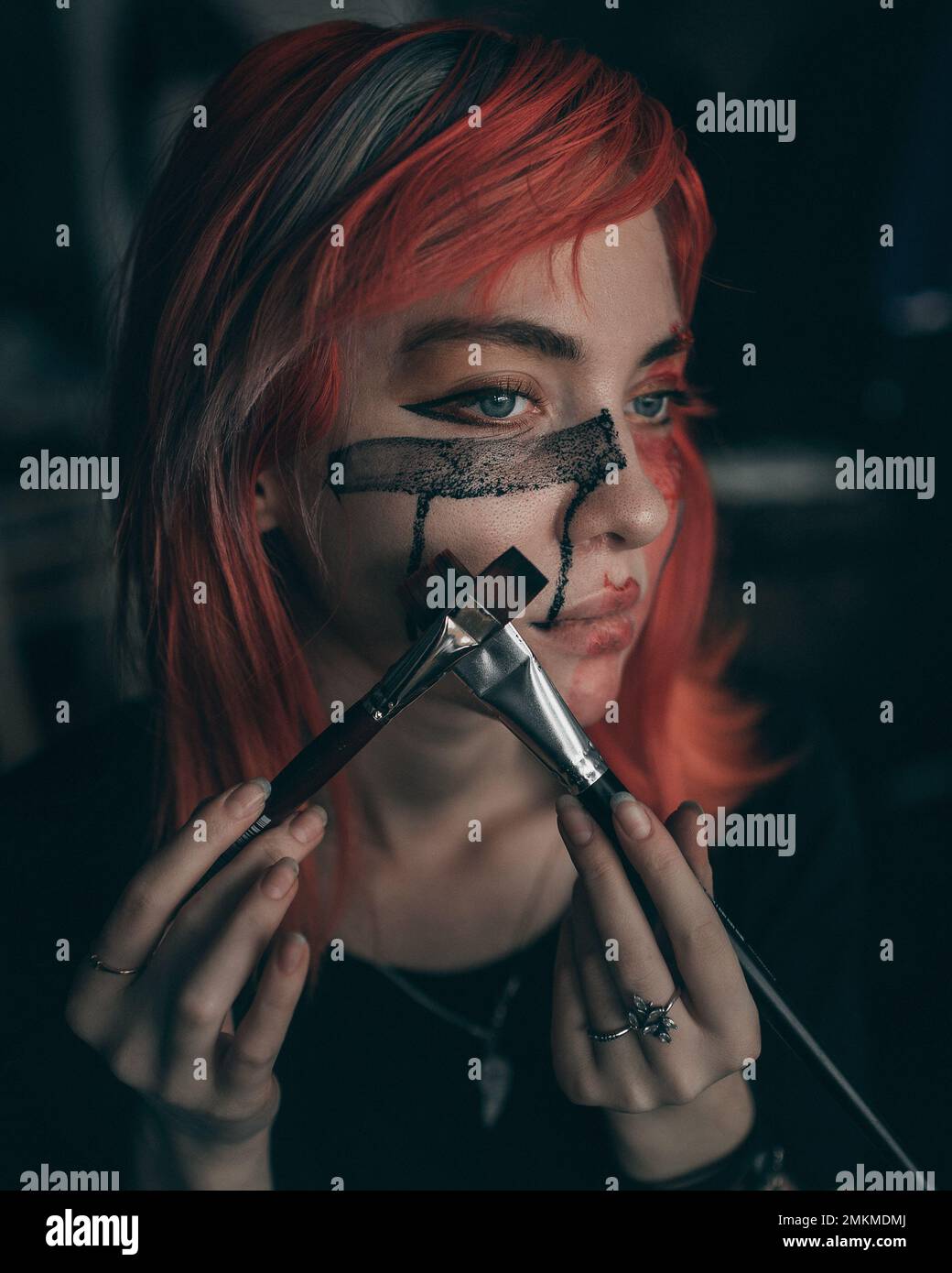 Young redhead woman artist applies red and black paint to her face with brush Stock Photo