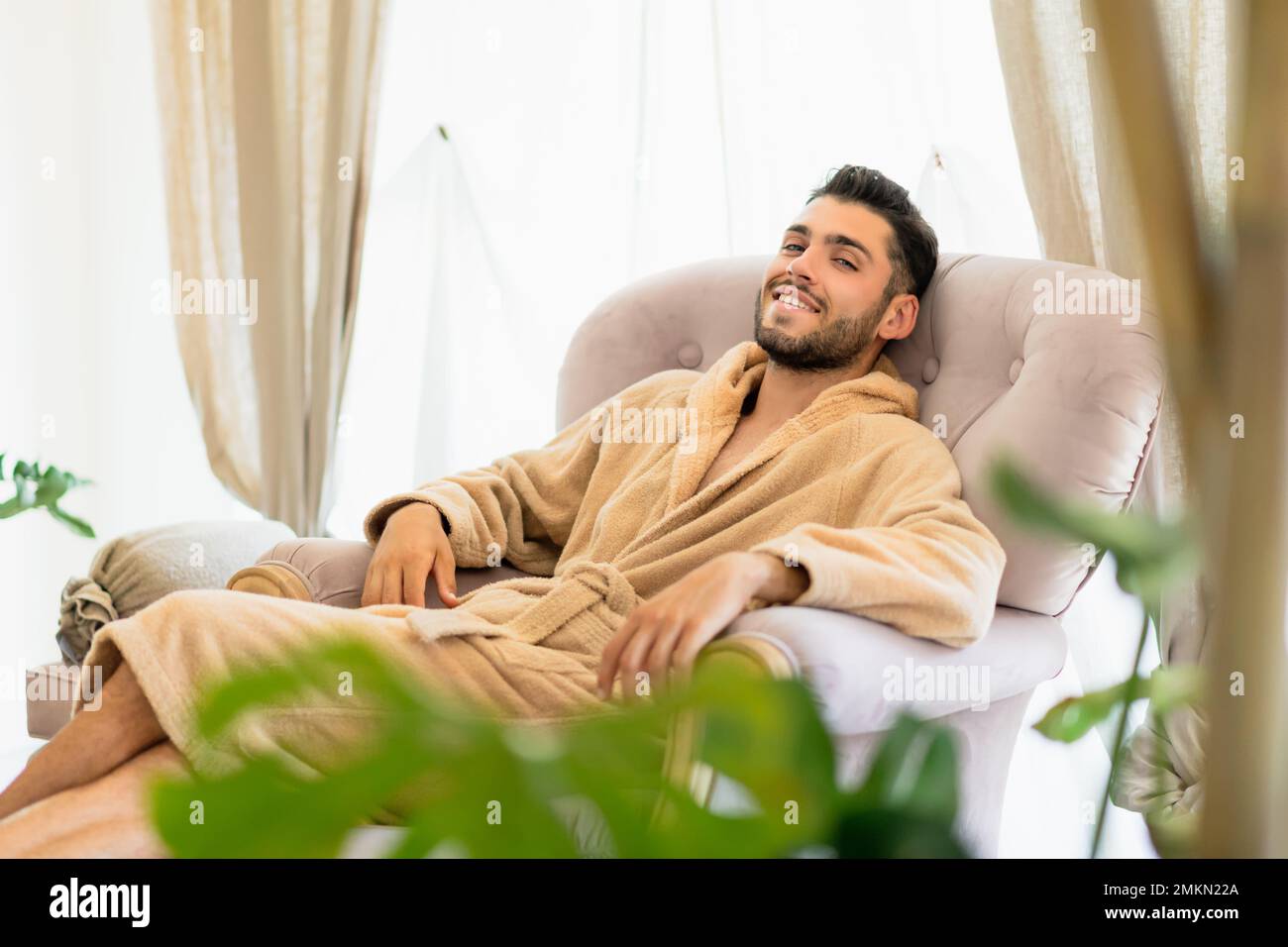 Young handsome bearded guy in soft bathrobe lounging in comfortable armchair in spa salon and looking at camera with smile Stock Photo