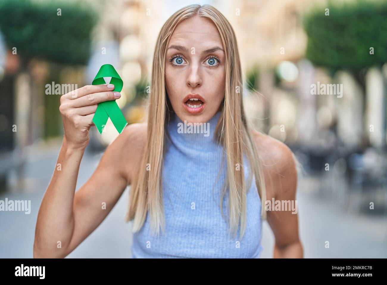 Young caucasian woman holding support green ribbon scared and amazed