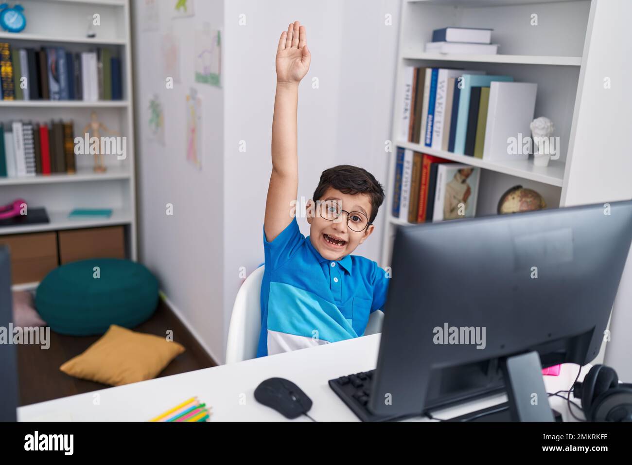 Adorable hispanic boy student using computer with hand raised up at ...