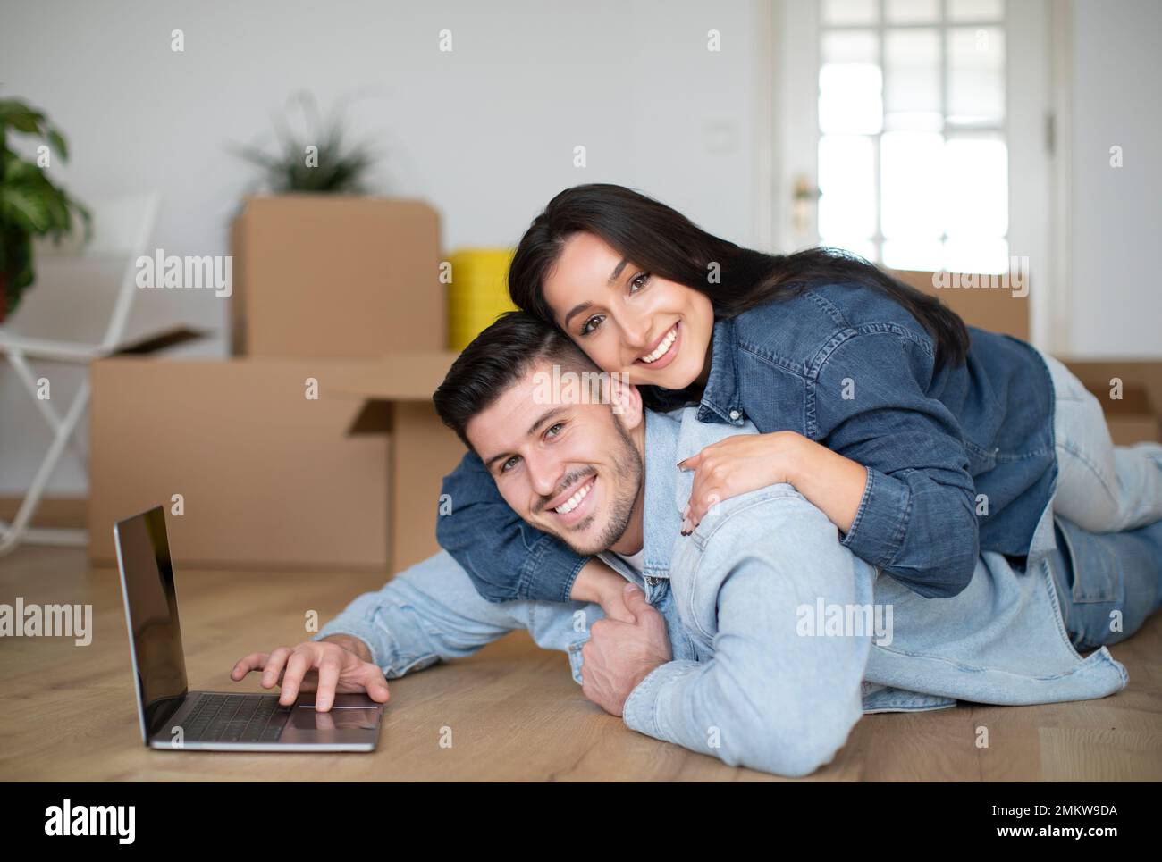 Happy Young Spouses Embracing And Using Laptop After Moving To Their New Home, Stock Photo