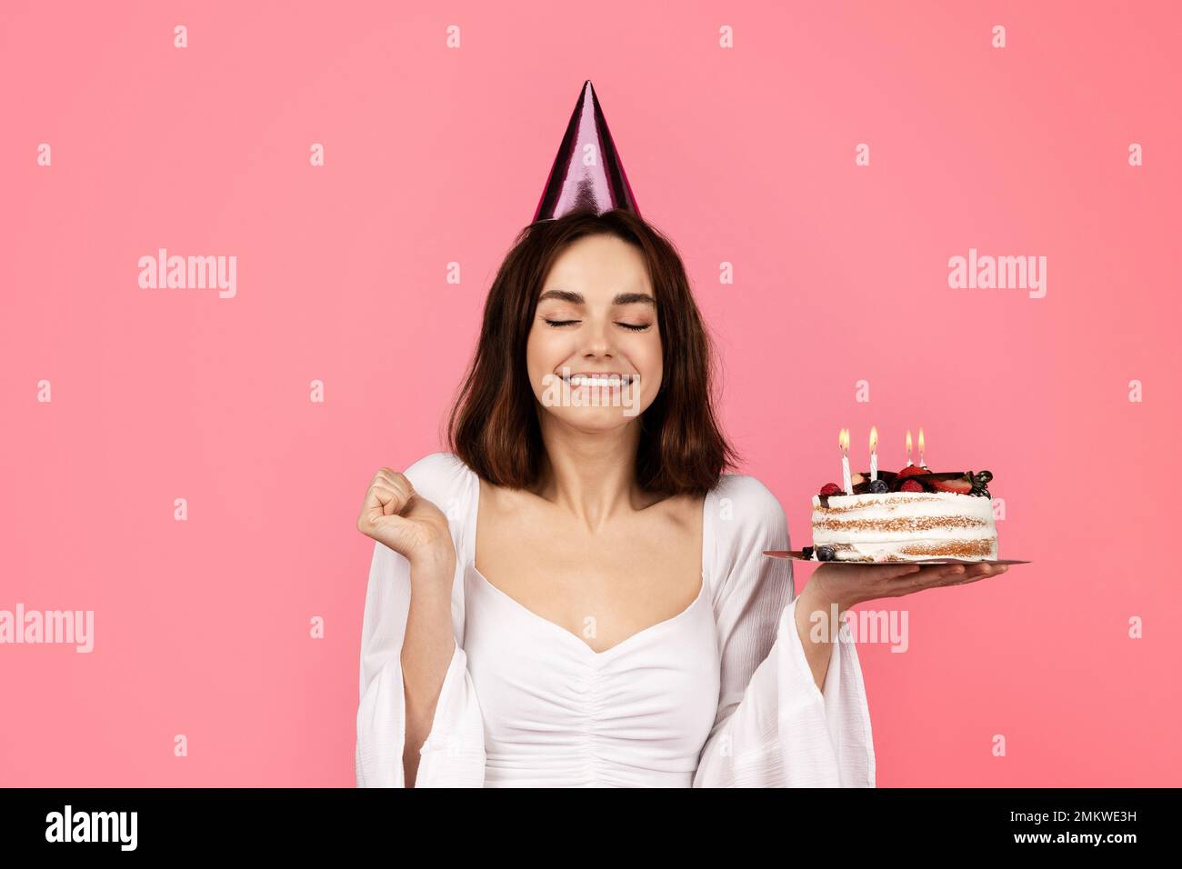 Cheerful young caucasian lady with closed eyes in hat hold cake with candles and dreaming, enjoy feast Stock Photo