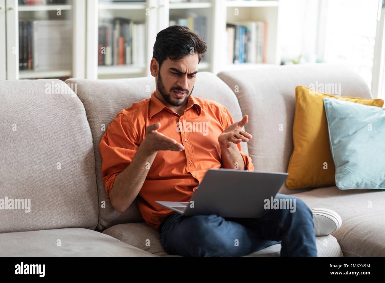 Irritated middle eastern man sitting on couch, using laptop Stock Photo