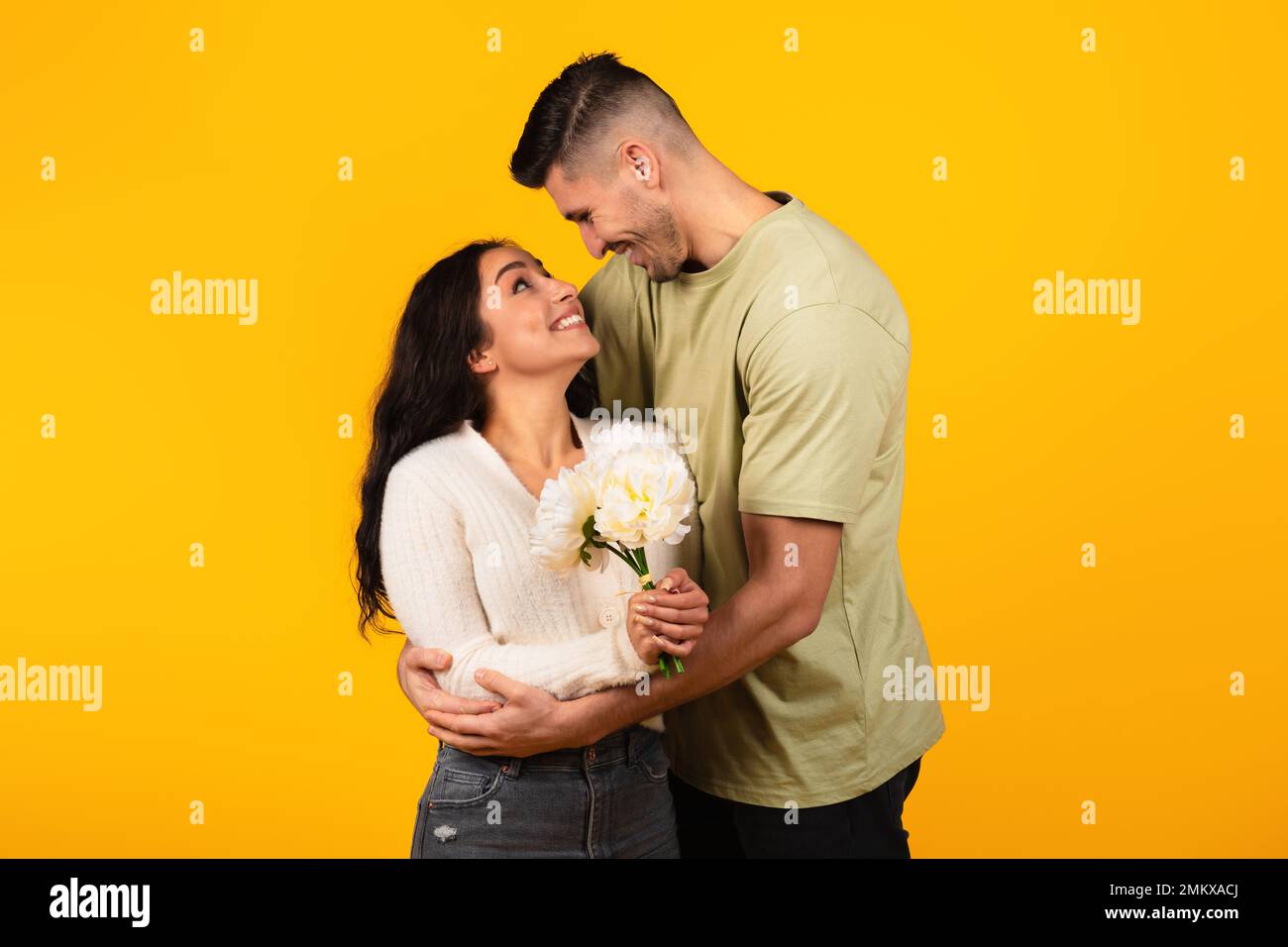 Cheerful young arab guy in casual hugs woman with flowers, enjoy romantic, isolated on yellow background Stock Photo