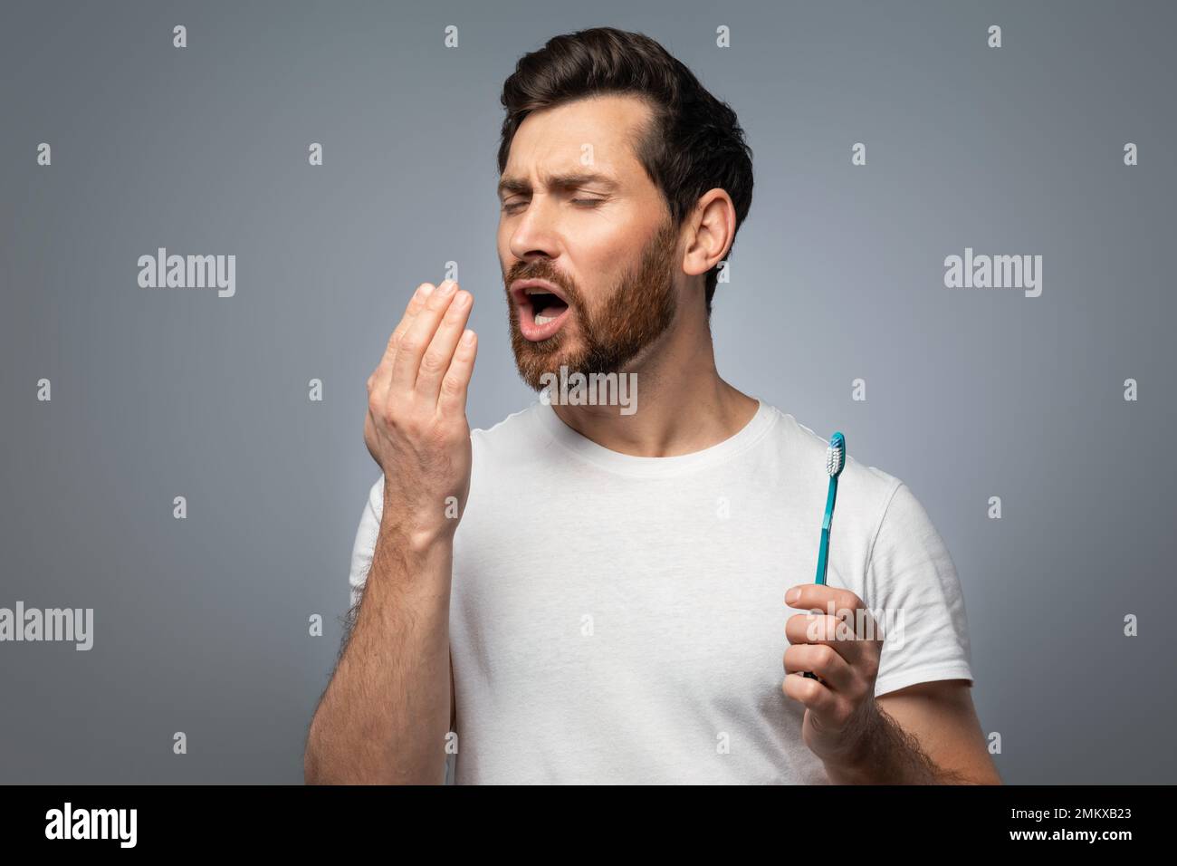 Bad breath. Handsome middle aged man checking his breath with his hand, blowing to it, standing over grey background Stock Photo