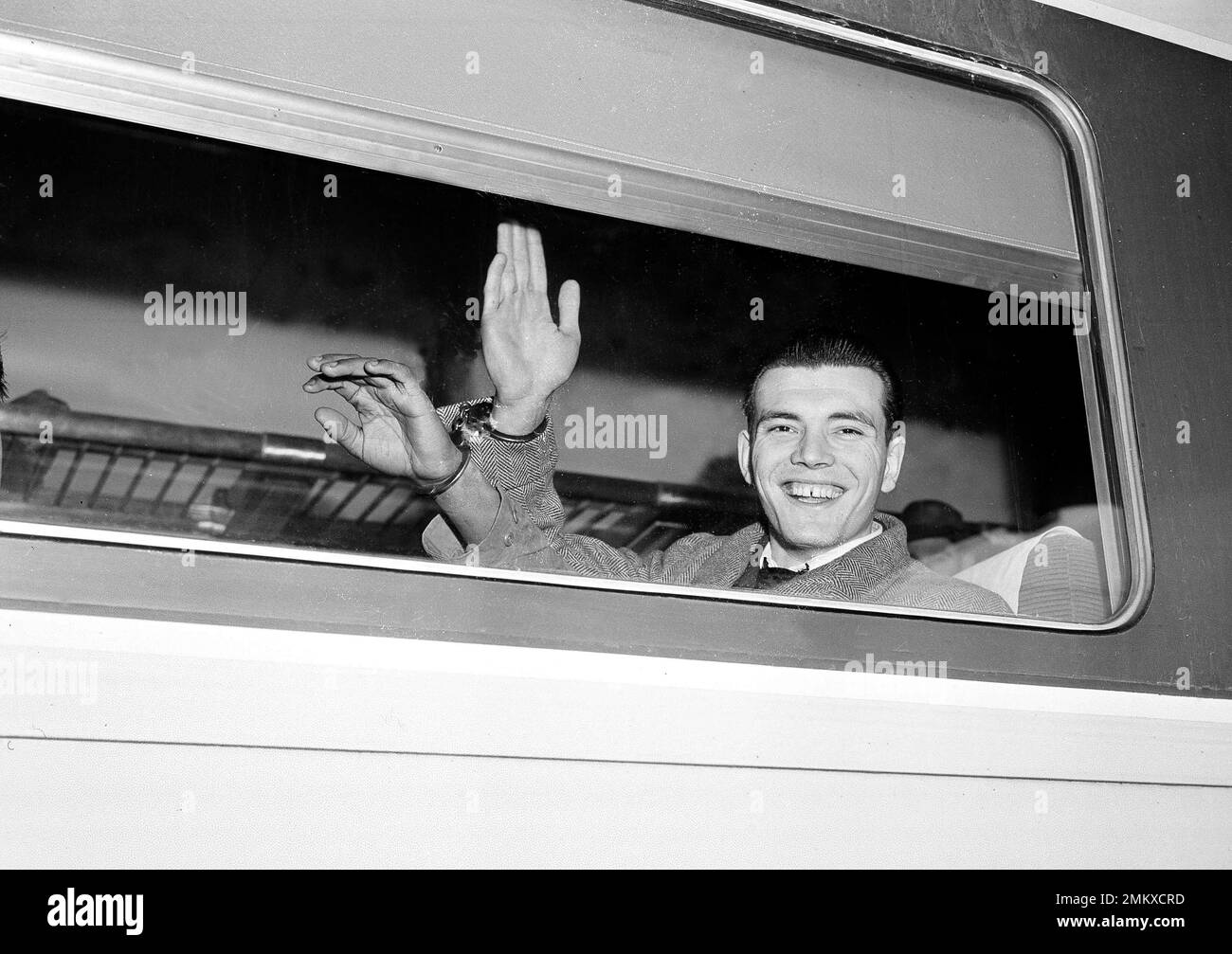 Ralph Edward Barrows, 20, formerly of Grand Rapids, Mich., smiles and waves his hand, which is handcuffed to that of another prisoner, in a train at Hoboken, N.J., March 7, 1950, as he leaves for the state prison at Elmira, N.Y. Barrows is under sentence from 20 to 40 years on a conviction of first degree manslaughter in the slaying of wealthy Canadian businessman, Colin Cameron MacKellar of Montreal. MacKellar was found dead in his Waldorf Astoria suite on Nov. 5, 1948. (AP Photo/Anthony Camerano) Stock Photo