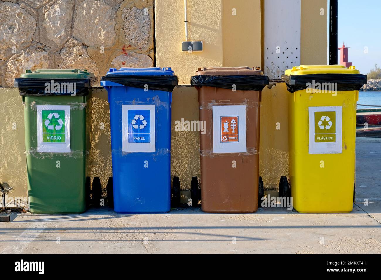 Four coloured wheelie bins in a row against a building. Each labeled with universal recycling symbols. Spanish words. Black wheels. Refuse bags. Stone Stock Photo