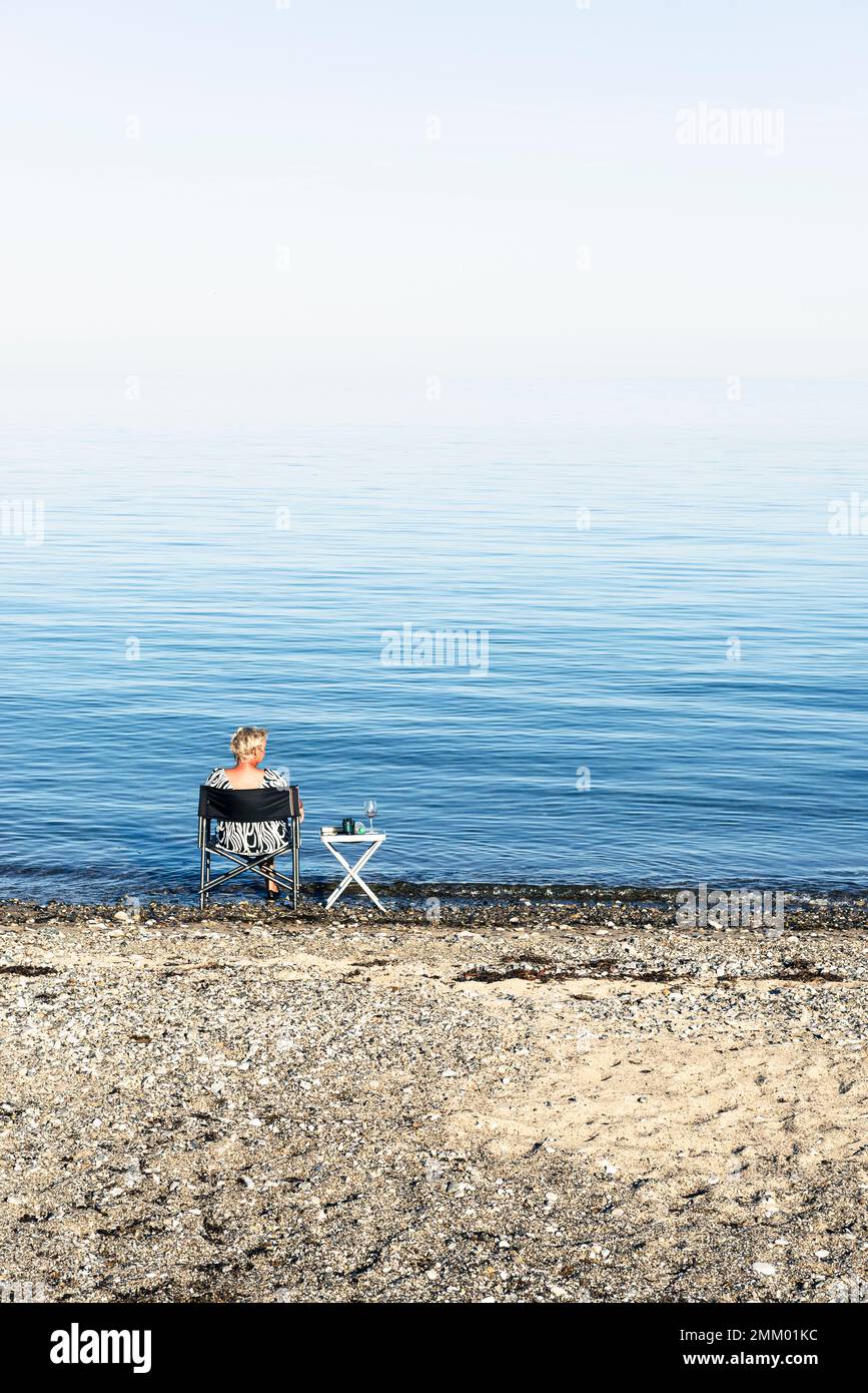 A blonde woman sits on a camping chair on the shore of Kattegatt in the sun and drinks wine, Djursland, Jutland, Denmark Stock Photo