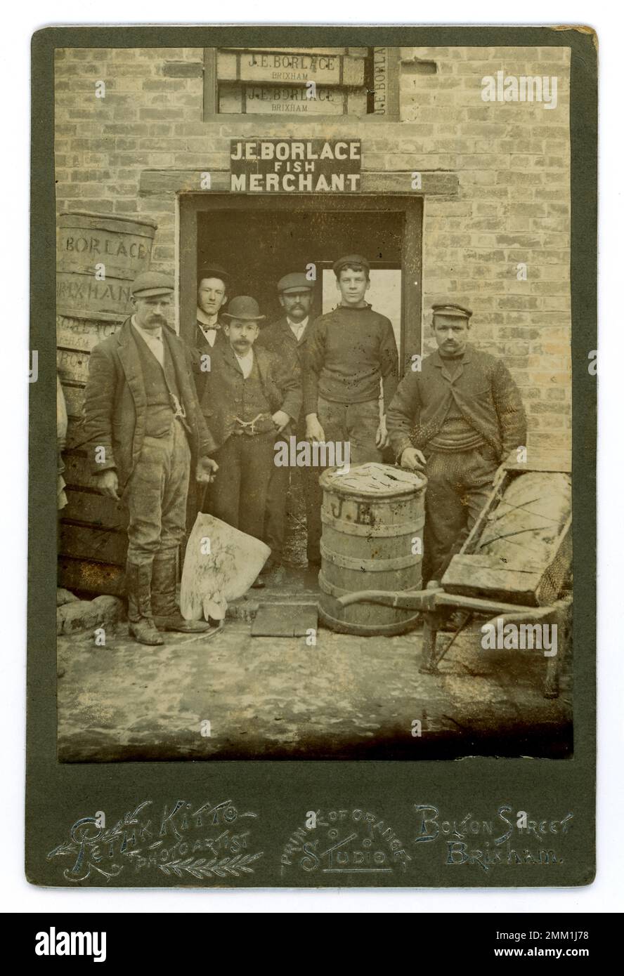 Original Victorian cabinet card cards of group of fishermen / traders / porters, characters / characterful,  outside the fish merchants of J.E. Borlace, with catch of fish, including a skate held up by one man, and a hand cart with wrapped produce, barrels advertising merchant's name, at the quayside, coastal fishing port of Brixham, Torbay district, Devon, U.K.  By photographer Peth K. Kitto (maybe Frederick K ) circa 1890's. Stock Photo