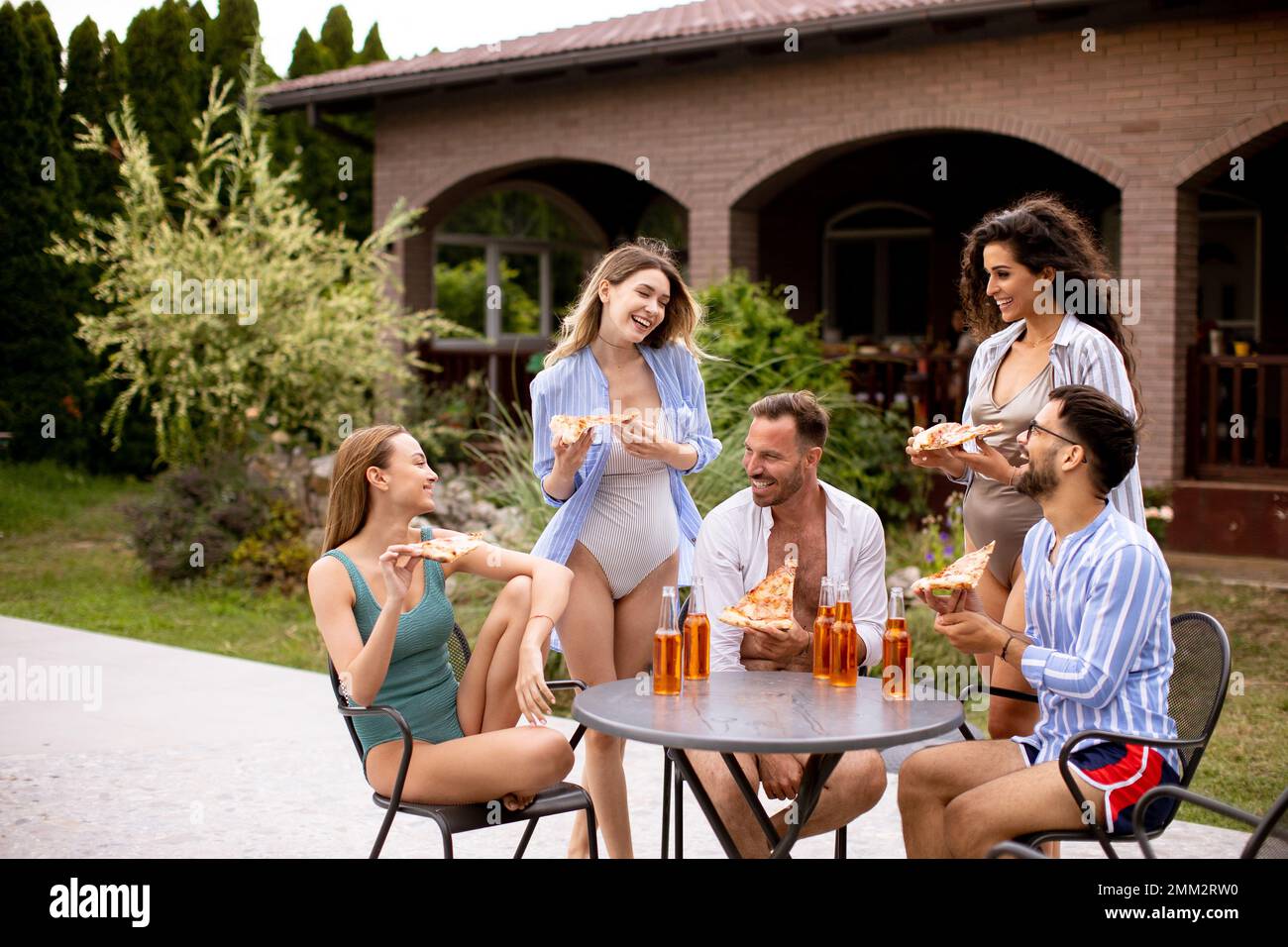 Group of happy young people cheering with cider and eating pizza by the pool in the garden Stock Photo
