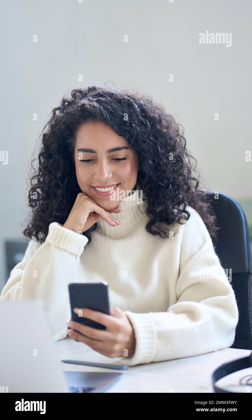 Smiling latin business woman using mobile phone in office sitting at desk. Stock Photo