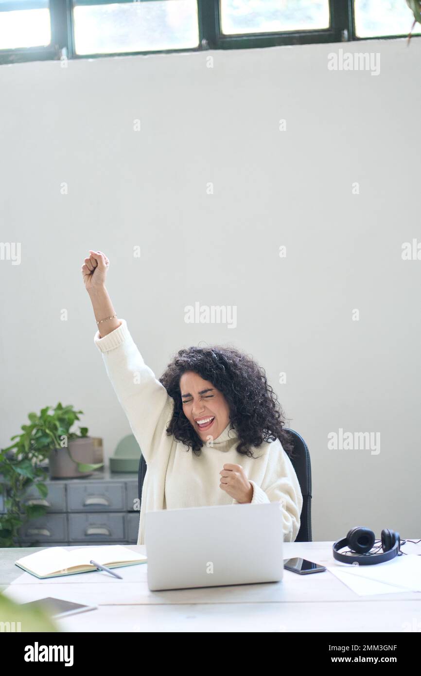Young happy latin woman student raising hand winning online looking at laptop. Stock Photo