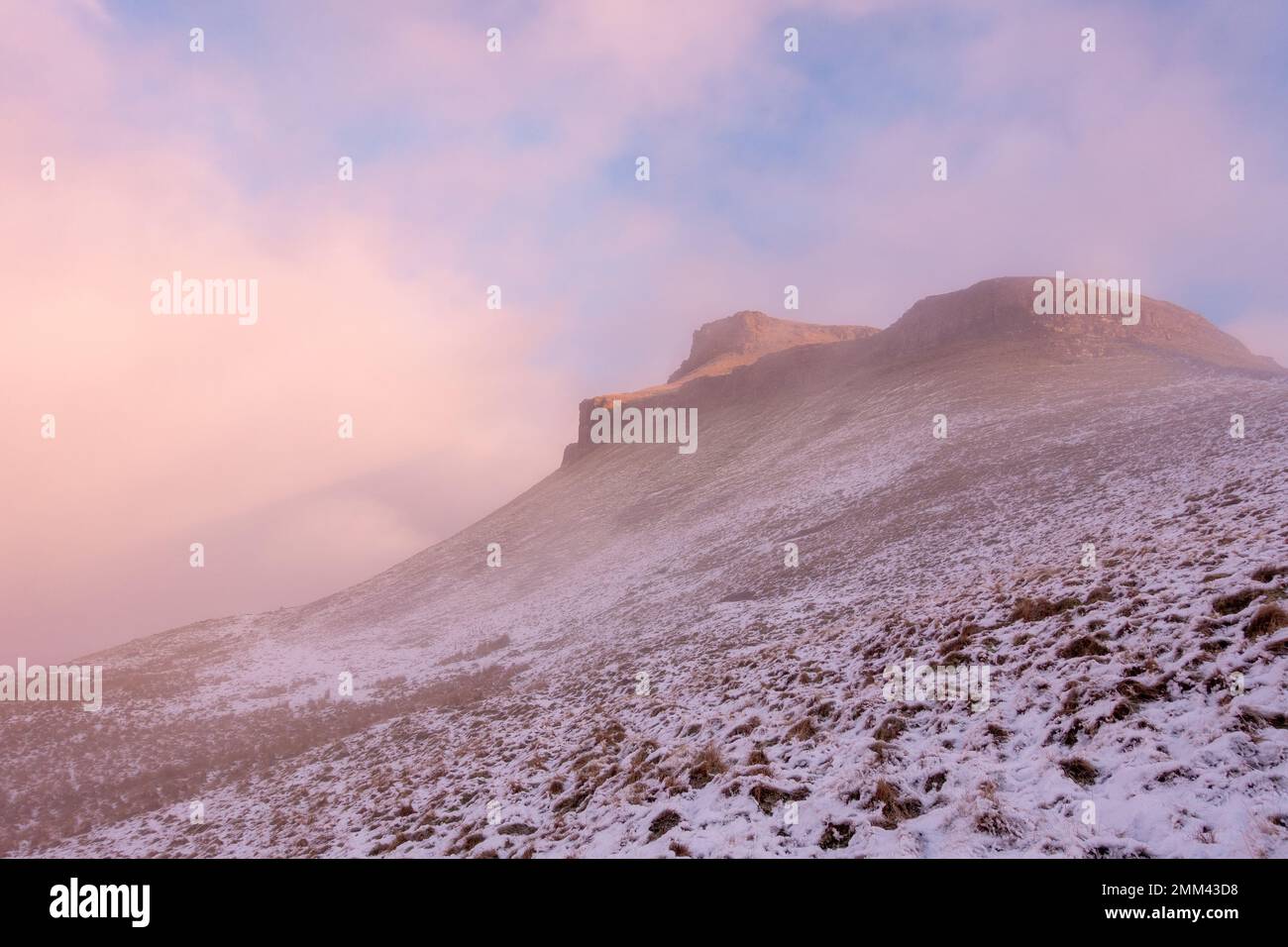 Looking up towards the summit of Pen-y-ghent mountain at sunrise in winter snow as the mist clears and the light catches the craggy top in the Yorkshi Stock Photo