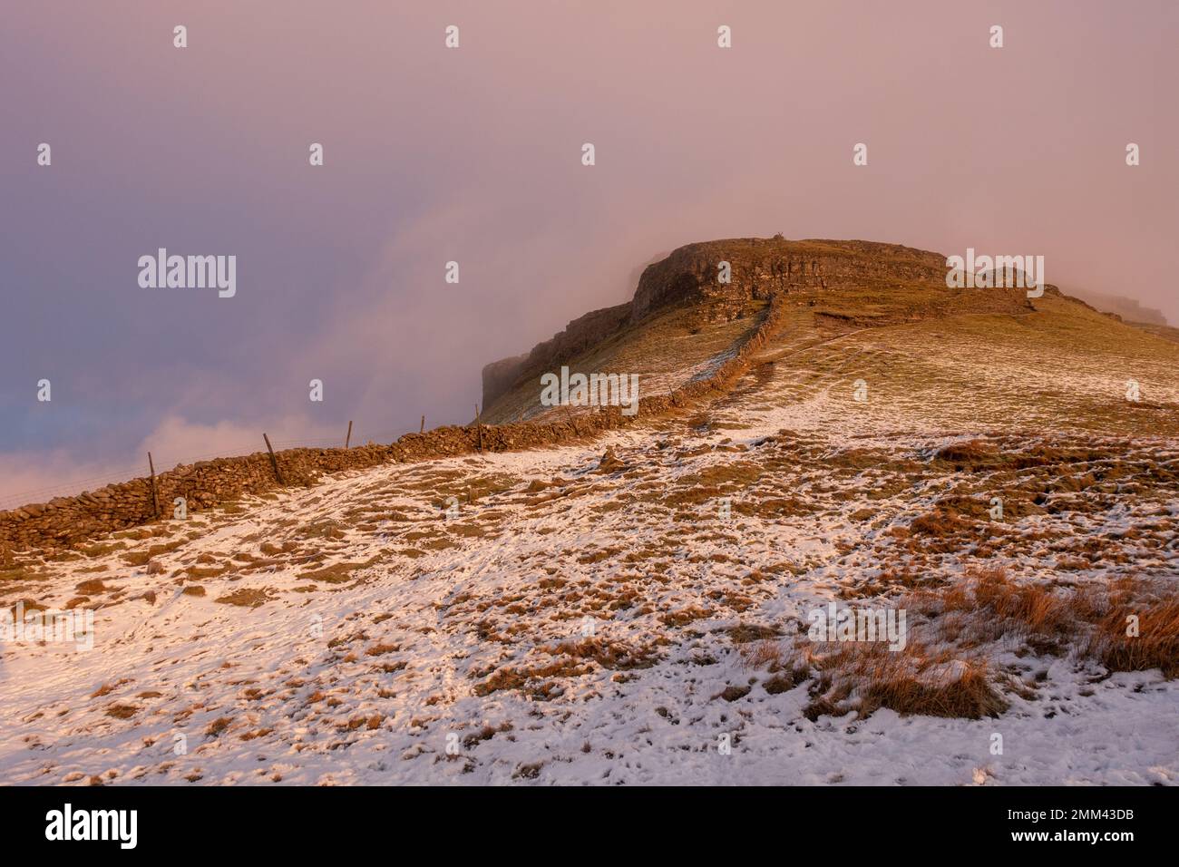 Looking up towards the summit of Pen-y-ghent mountain at sunrise as the mist clears and the light catches the craggy top in a snowy Yorkshire Dales Na Stock Photo