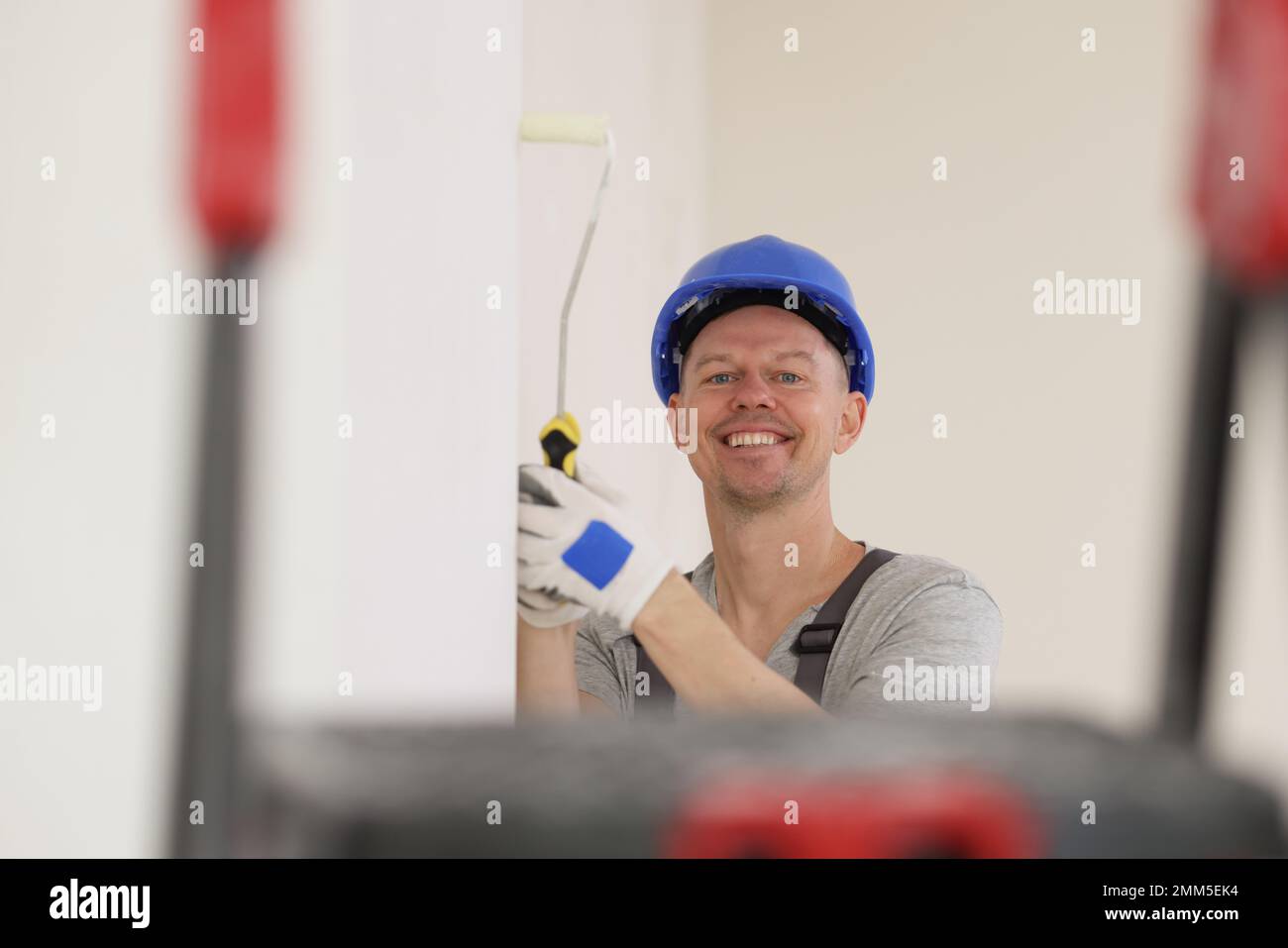 Happy painter in hard hat paints wall with white paint and looks at camera. Stock Photo