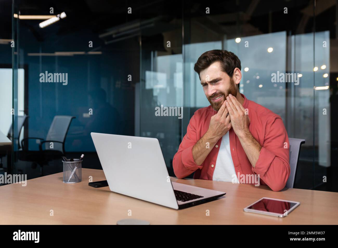 A young man, businessman, freelancer, designer sits in the office at the table. works on a laptop. He holds his cheek, feels discomfort in his mouth, has a severe toothache. Stock Photo