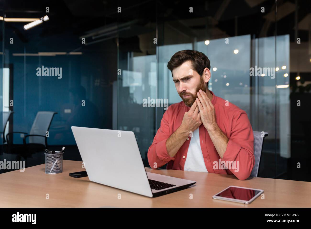 A young man, a student, a freelancer sits at the desk in the office. studies, works on a laptop. He holds his cheek, has a severe toothache. Stock Photo