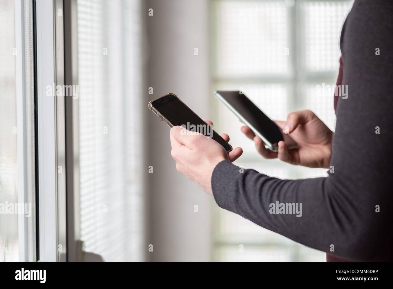 Close up of a man holding two smartphones. Man using two phones at the same time.Busy man standing next to windows and using two smartphones. Stock Photo