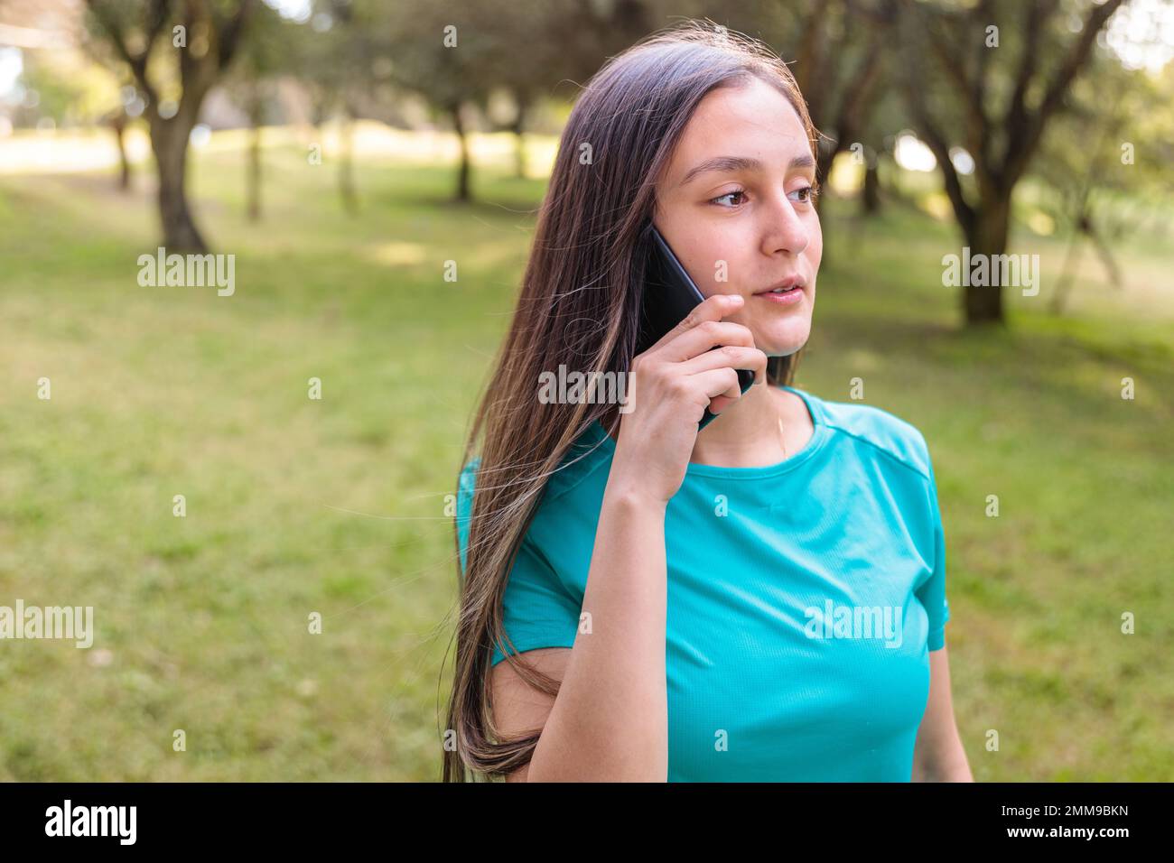 Teenage student girl wearing aquamarine t shirt, making a phone call to her family in the park. Sun backlight Stock Photo