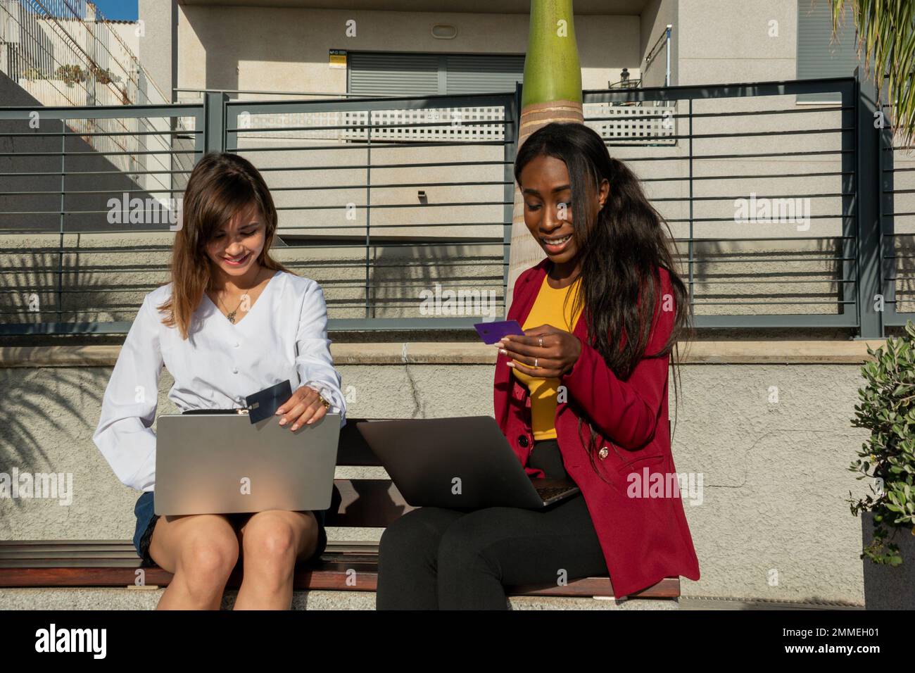 Two smiling businesswomen sitting on a bench outdoor shop online with ...