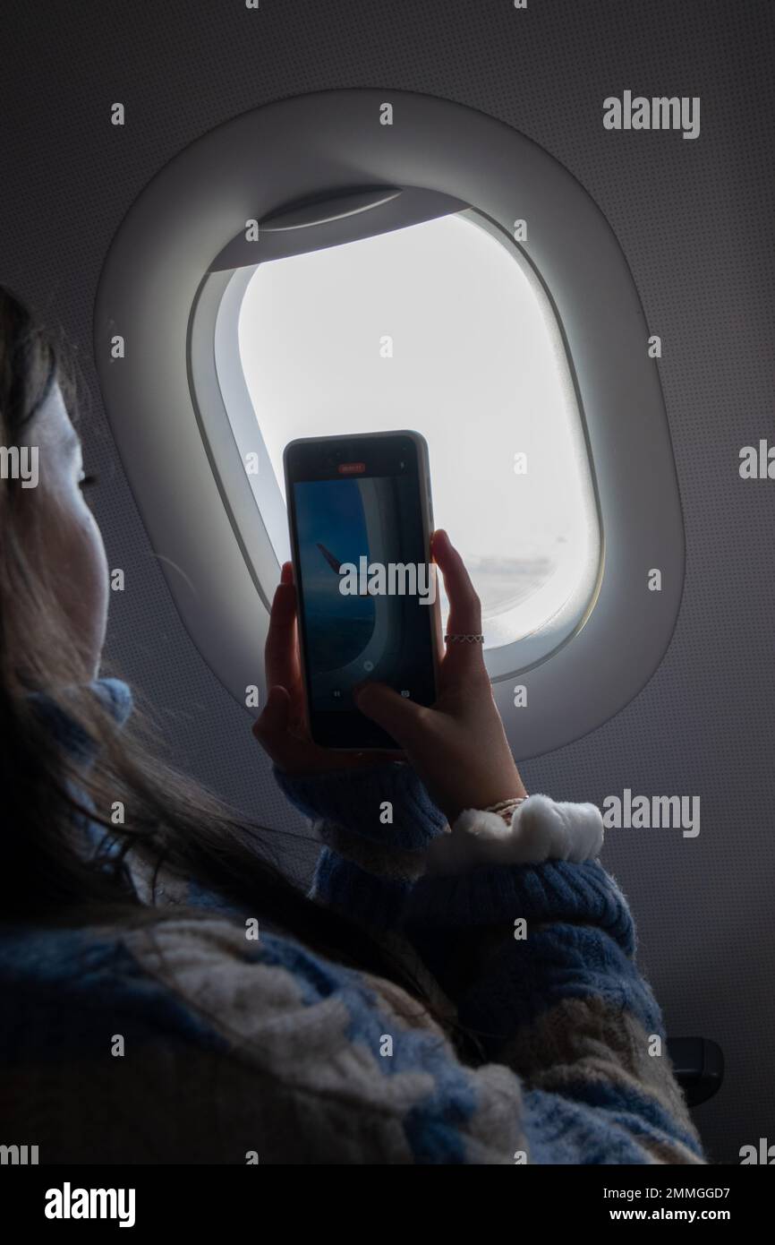 Young girl sitting at the window seat on airplane while taking pictures when flying in the sky. Kids traveling on airplanes. Teenagers passengers. Stock Photo