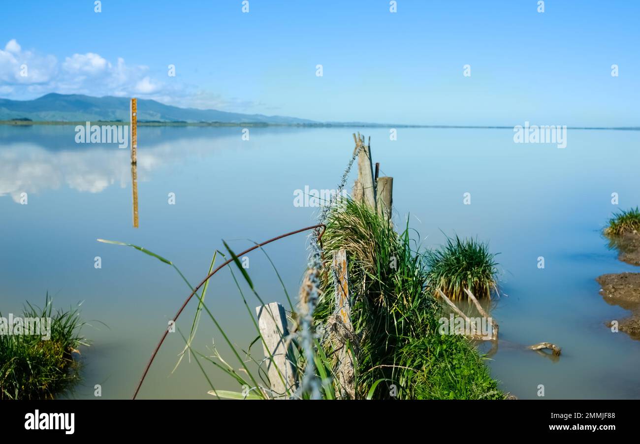 Fence leading into calm blue water of Lake Wairarapa with distant hills across other side in New Zealand North Island. Stock Photo